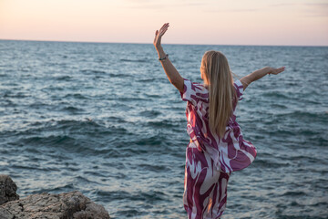 blonde woman on a rock by the sea at sunset hands up