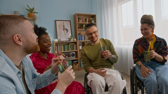 Joyful company of young multiethnic friends and wheelchair users toasting and clinking lemonade glasses, laughing and chatting while hanging out at home in cozy living room