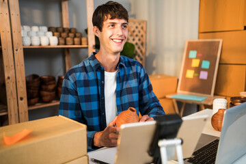 Smiling young male entrepreneur showcases handmade ceramic pottery while live streaming from his small business workspace. Promoting craft products through online marketing and e-commerce.