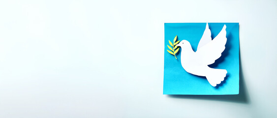 Obraz premium A white paper dove cutout holding a green olive branch on a blue square background. The minimalist design evokes themes of peace, hope, and reconciliation, ideal for symbolic or cultural projects.