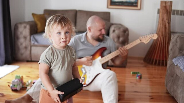 An attractive father playing guitar to his adorable baby boy in living room. Male kid listening and smiling. Concept of family, childhood, relationship and parenthood. Music songs writing. Cild dad