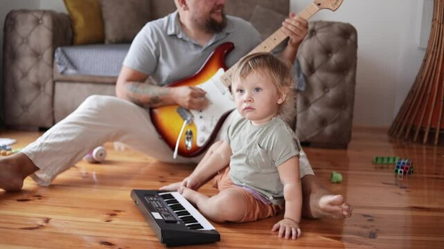 An attractive father playing guitar to his adorable baby boy in living room. Male kid listening and smiling. Concept of family, childhood, relationship and parenthood. Music songs writing. Cild dad