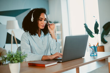 Young woman in casual wear with headphones working on laptop in stylish home office space during the day