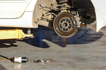 The vehicle lifted from the hydraulic lift shows brake discs and suspension, the rear wheels have been removed for maintenance, and tools are placed on the ground nearby.