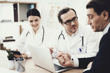 Doctor in glasses advises patient sitting at table with laptop in medical office.