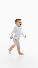 Joyful little boy walking barefoot in white shirt and beige shorts over white background. Concept of childhood, movement, joy, health, growth, energy, physical development.