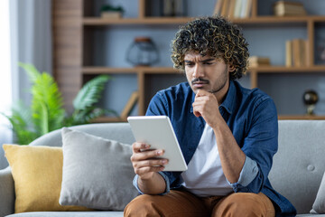 Serious thinking man using tablet computer reading e-book while sitting on sofa in living room at home.