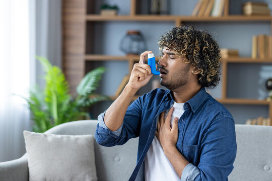A young man with curly hair is using an inhaler for his asthma while sitting on a sofa in a bright living room.