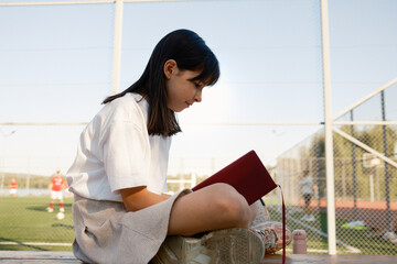 Side profile of a young girl absorbed in a book while sitting near a sports field. The scene...
