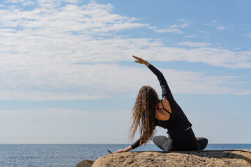 A woman doing yoga on  a rocky shore the ocean. She wears black yoga attire, embracing mindfulness and tranquility. The peaceful sea and sky create a calming atmosphere.