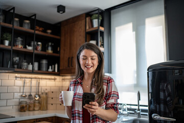 Smiling woman using smartphone and drinking coffee in kitchen
