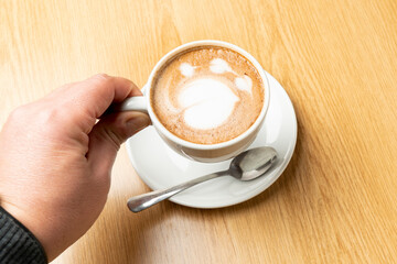 Hand holding a white cup of cappuccino with latte art on wooden table, in a cafe or restaurant