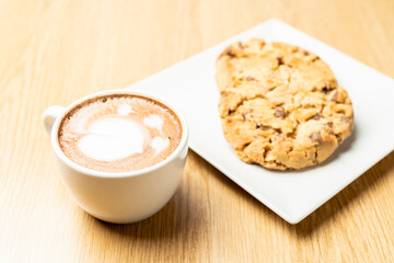 Two chocolate chip cookies on a white plate beside a cup of cappuccino with latte art, placed on a wooden table, creating a cozy and inviting atmosphere.