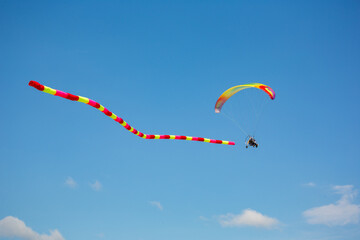 A powered paraglider flying in the blue sky with a long, colorful windsock trailing behind