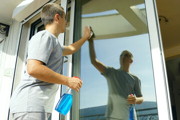 Fototapeta premium Cleaning of office premises: a man washes a glass door with a cloth and glass cleaner in a spray bottle. Lowering social status as a result of moving to another country: a migrant works as a cleaner