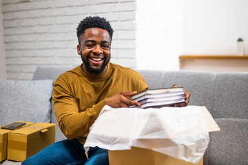 Happy young African American man smiling and opening delivered package with books while sitting on...