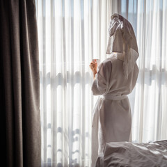 Beautiful young woman in a white bathrobe with a towel on her head stands near the window.