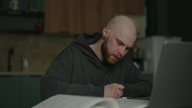 Focused pensive male student researching reading book, taking notes on notebook sitting at kitchen table with laptop and textbook in apartment, focusing intently during late night study session.