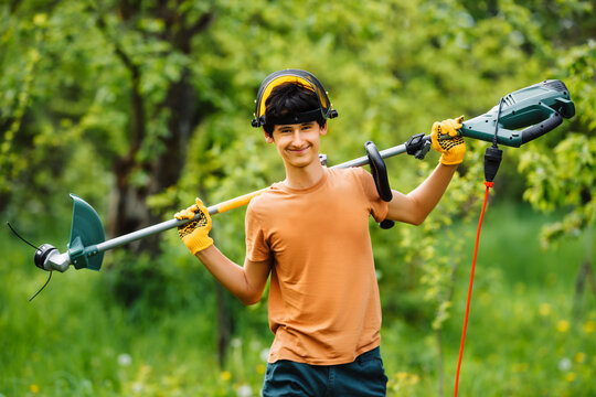 Young smiling teenage boy gardener posing in the garden holding an electric scythe.