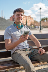 Young man sitting on bench using smartphone and headphones in city park © Alejandro