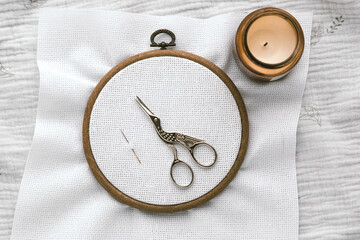 Embroidery hoop with white canvas, vintage stork-shaped scissors and needle on textured fabric, next to a lit candle in warm cozy atmosphere, representing creativity and handmade crafts, cross stitch © Evgeniia Primavera