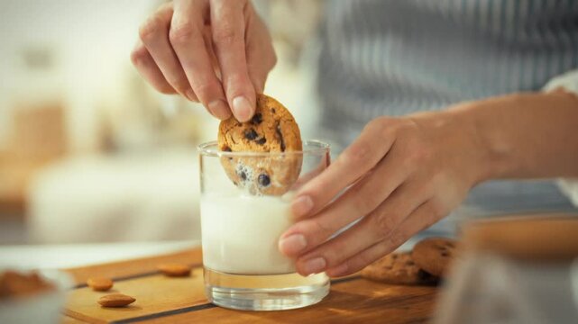A person dips a chocolate chip cookie into a glass of milk in a bright kitchen. The atmosphere is warm and inviting, suggesting a moment of comfort and enjoyment