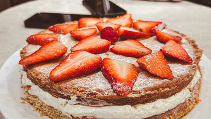Leicester, UK. 10 November 2024. A freshly baked Victoria sponge cake topped with sliced strawberries and dusted with icing sugar, filled with whipped cream and jam, served on a kitchen counter.
