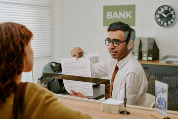Obraz premium Businessman in professional attire sitting in bank office discussing documents with customer at bank, office environment with visible bank sign and clock in background