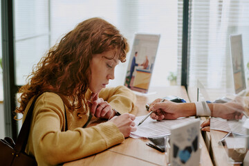 Woman sitting at office desk reviewing documents while colleague points at paper, scene inside a...