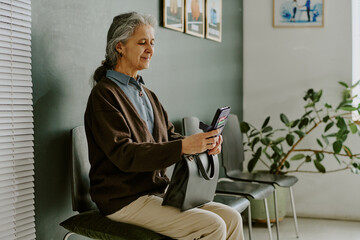 Elderly woman sitting on a chair in a modern room, checking her smartphone and holding a handbag, with framed photos and a plant in background