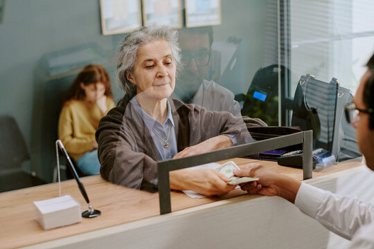 Senior woman receiving money from bank teller seated behind counter, modern office setting with other people engaging in various activities in background