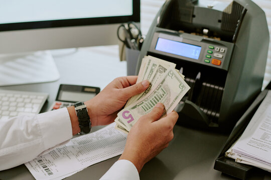 Person counting cash using a currency machine at office workspace with various tools and devices, displaying financial transactions and business environment