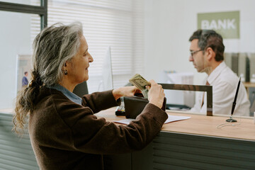 Senior woman handing money to bank teller at service desk during transaction interaction, bank...