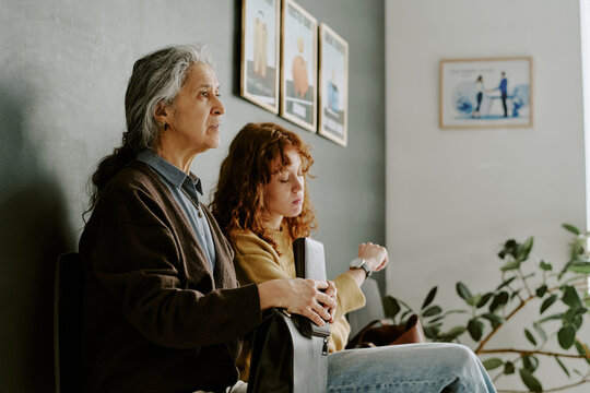 Elderly man with long hair sitting near young woman in office lobby, both waiting with calm expressions surrounded by artwork on wall, plant in background