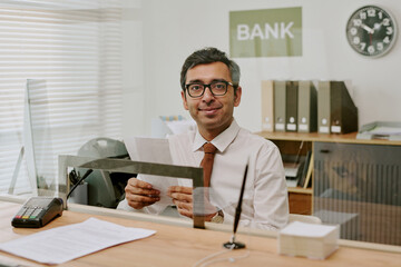 Portrait of smiling banker in glasses and tie holding papers and sitting at desk in office with files and documents in background