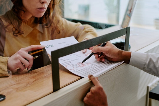 Detailed view of person filling out forms at bank counter, interacting with bank teller behind glass divider in background