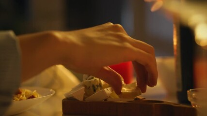 A hand delicately selects gourmet cheese from a wooden board during an evening gathering. The warm light creates an inviting atmosphere as guests savor delicious appetizers and share moments