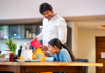 Indian Father lovingly prepares tiffin and places it in school bag while daughter eats breakfast