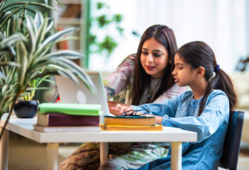 Mother sits beside her daughter as she attends an online class on a laptop, encouraging learning