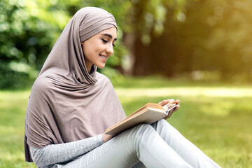 Relaxed muslim woman enjoying weekend at park, sitting on grass and reading book, empty space