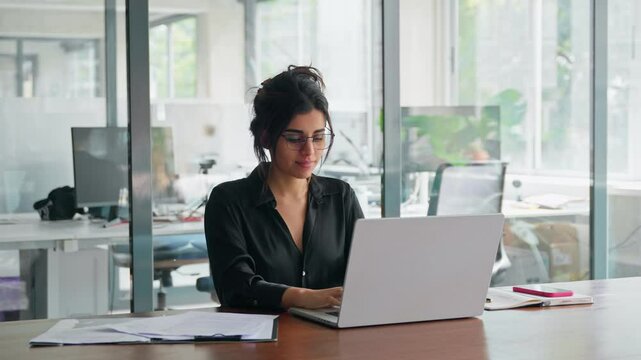 Young professional it specialist latin hispanic business lady working on laptop pc sitting at desk in modern office space. 30s middle eastern indian woman using computer technology app for work online