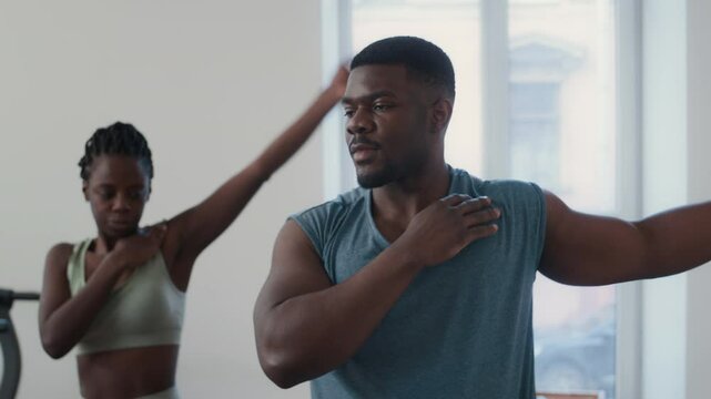 Black fitness trainer demonstrating arm circle exercise to young woman while leading pre-workout warmup in gym