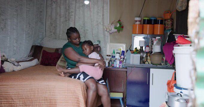 Poverty. Inequality.  Wide Panning. Black African mother and young daughter sitting on a bed in their small room shack in a township in Africa
