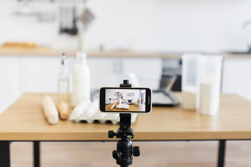 Modern kitchen setup with smartphone capturing scene of cooking ingredients like flour, eggs, milk. Perfectly organized workspace for creating recipes or food-related video tutorials.