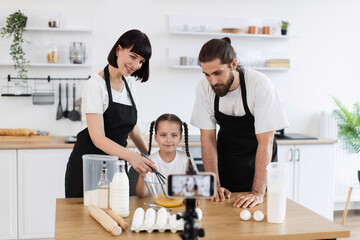 Caucasian family of bloggers filming cooking tutorial. Woman, man, and young girl preparing recipe, kneading dough in modern, bright kitchen environment while camera records.