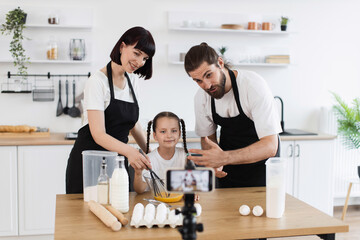 Obraz premium Caucasian family of three, parents and child, creating culinary content in bright setting. Woman, man, and girl filming cooking process with whisk and ingredients, wearing aprons