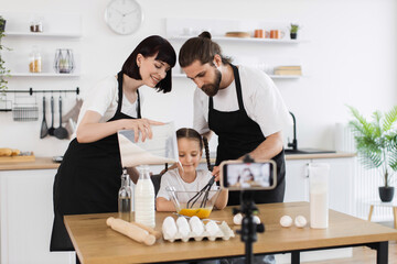 Obraz premium Caucasian family of three, adult parents and child, recording baking tutorial in home kitchen. Father, mother, daughter bonding through learning culinary skills