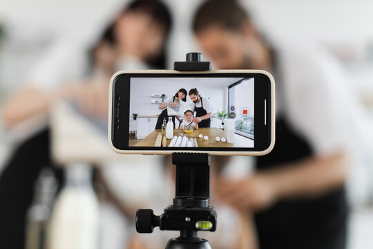 Caucasian family of food bloggers recording baking tutorial in modern kitchen with smartphone. Parents and child knead dough with whisk bowl while shooting creative content