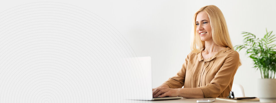 A professional woman with long blonde hair is smiling while typing on her laptop. She sits at a modern desk in a well lit office space, surrounded by a green plant and a mobile device, copy space