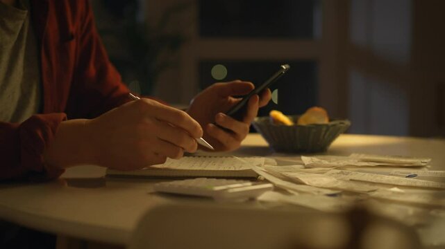 A man, sitting at a table in the kitchen, painstakingly calculates expenses in the evening, looks through receipts and enters data into a calculator. Warm stage lighting, comfortable view